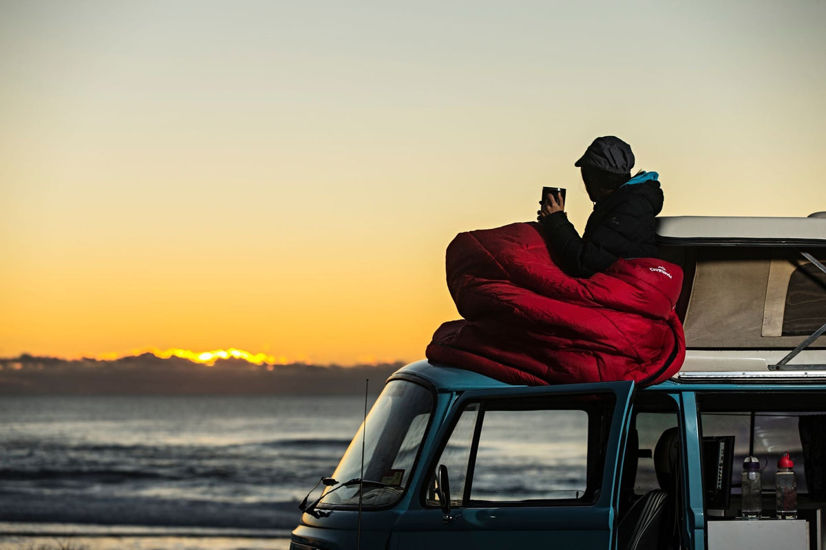 A camper is watching the sunset wrapped in his sleeping bag.